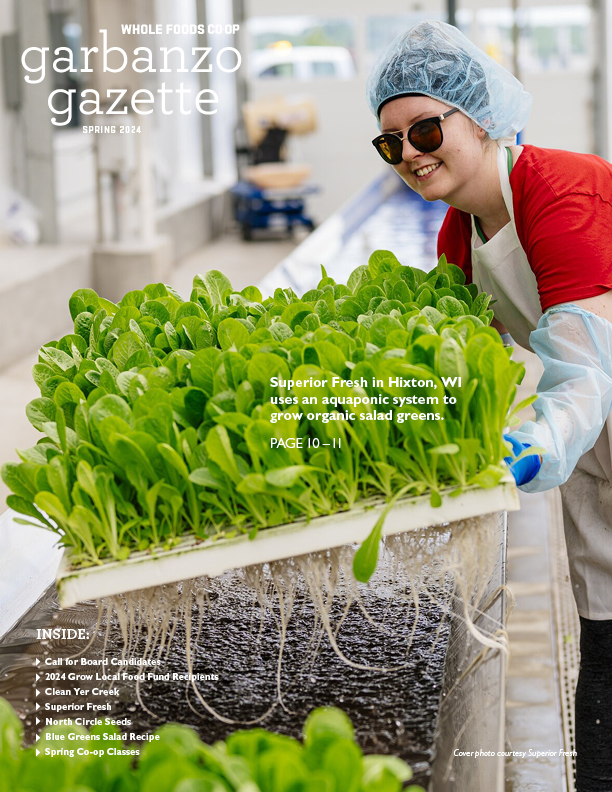 A person wearing a hairnet, safety glasses, and gloves smiles while holding fresh leafy greens from an aquaponic system. The cover text reads “Garbanzo Gazette - Spring 2024” and highlights include “Superior Fresh in Hixton, WI grows organic salad greens” and other articles from Whole Foods Coop Duluth MN.
