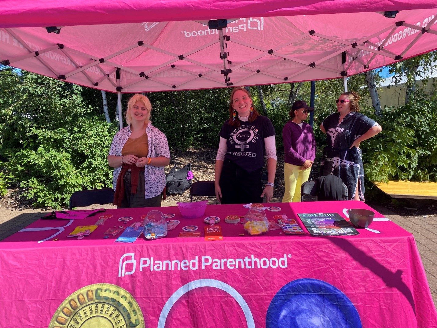 Two people are standing behind a Planned Parenthood booth covered with a pink tablecloth displaying various informational and promotional materials. The booth is under a pink canopy with the Planned Parenthood logo, set against a backdrop of greenery and a few other people.