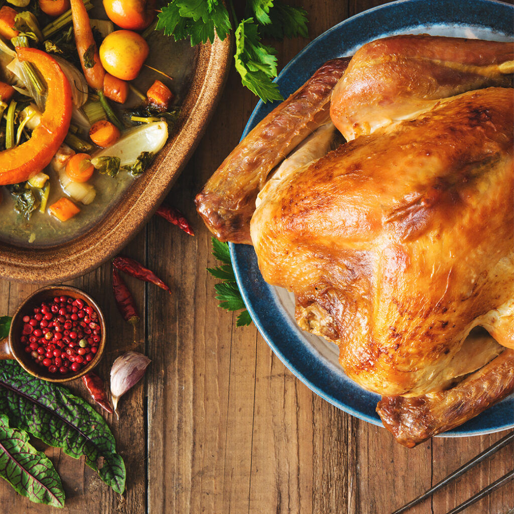 A roasted whole turkey on a blue plate is placed on a wooden table, surrounded by various vegetables, including pumpkins, carrots, and greens. Thanksgiving spices and herbs, such as red peppercorns, garlic, and chili peppers, are also arranged around the table.