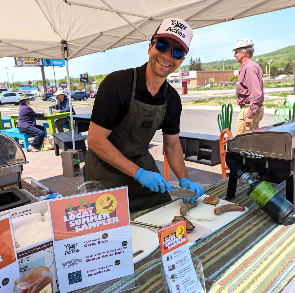 A smiling person in a black apron, cap, and gloves is slicing sausages at an outdoor food stall reminiscent of the lively atmosphere at Whole Foods Coop in Duluth, MN. The stall, set under a tent, features various food items including garlic bratwursts. Other people and a scenic outdoor background are visible.