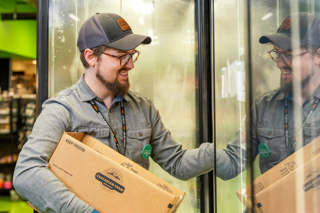 A person with glasses, a beard, and a cap is smiling while holding a cardboard box labeled 