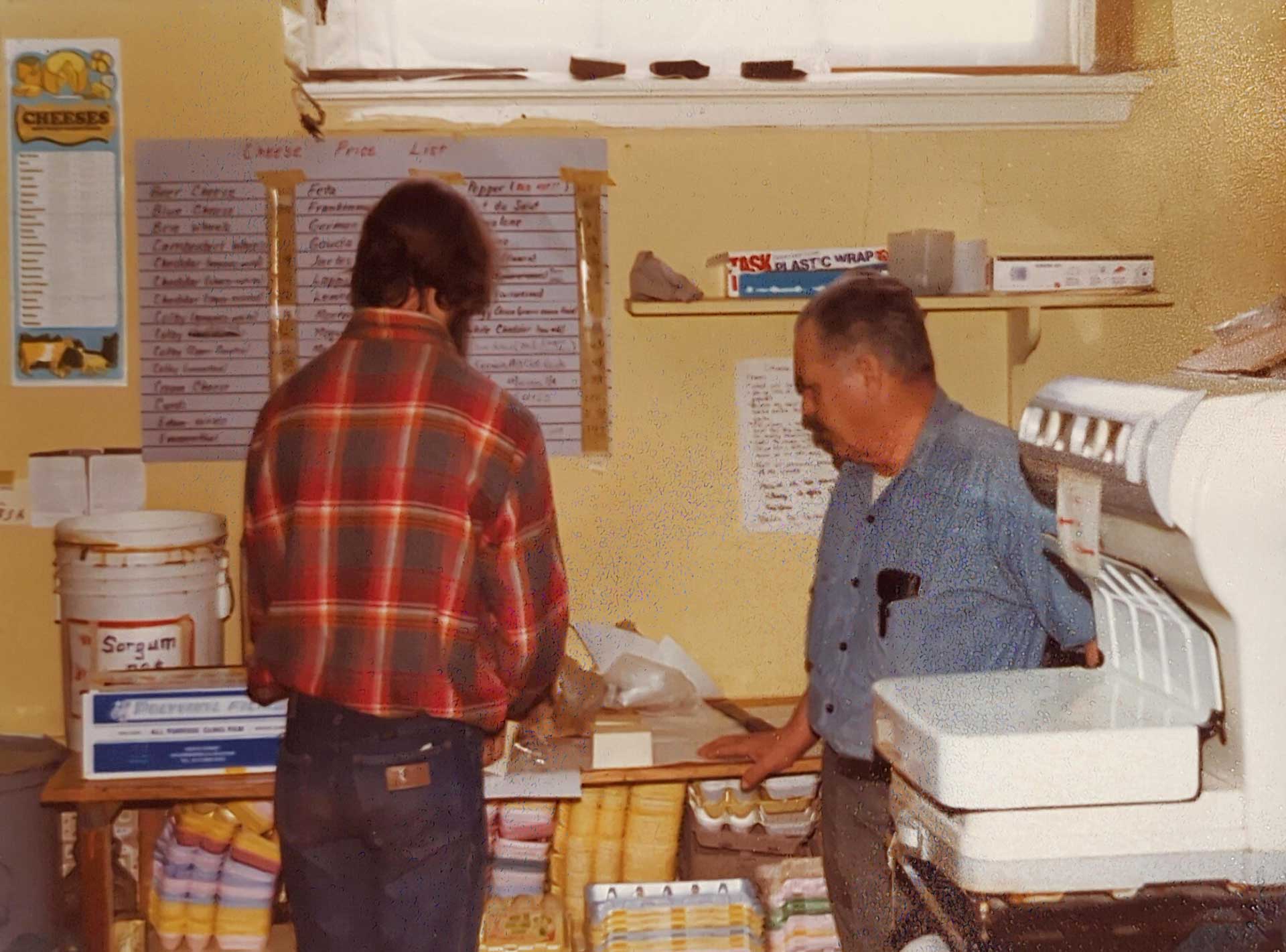 Two men stand in a small shop. One man in a red plaid shirt faces away, examining items on a counter. The other man in a blue shirt faces left, looking at the counter. Shelves display various products, reminiscent of those found at Whole Foods Coop Duluth MN, and a menu hangs on the wall above them.