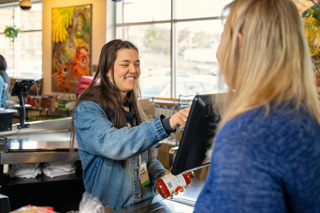 A cashier wearing a denim jacket scans items at the checkout counter while smiling and chatting with a female customer. The counter holds various products, and the background features vibrant artwork and a large window revealing an outdoor scene.