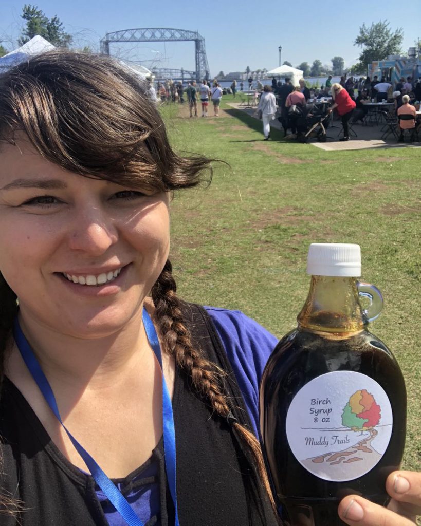 A person smiles while holding a bottle of birch syrup labeled 
