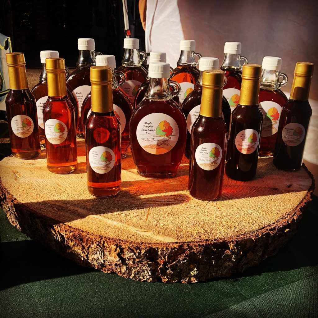 A variety of bottles filled with amber liquids are arranged on a large, circular wooden slab. The bottles have white labels featuring a leaf design. They are displayed in a well-lit setting, possibly an outdoor market or fair.
