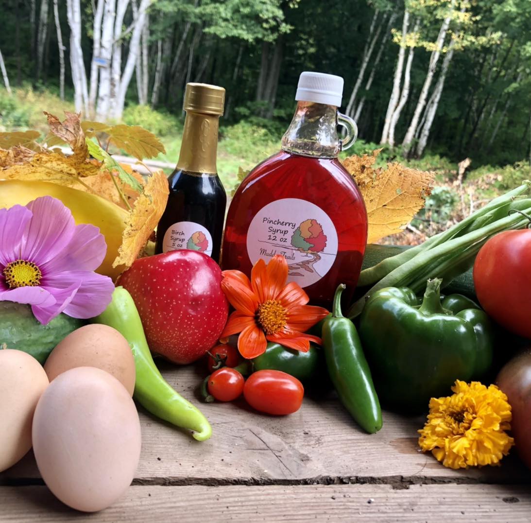 A display of farm produce includes a bottle of syrup, a smaller bottle of another liquid, an array of fresh vegetables and fruits, and a few flowers. Eggs, tomatoes, peppers, and apples are visible, all arranged on a wooden surface with a forest in the background.