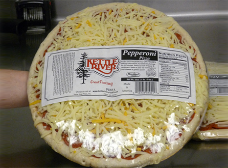 A person is holding an uncooked Kettle River pizza, topped generously with shredded cheese and pepperoni. The visible label lists nutrition facts and ingredients. In the background, a kitchen counter displays various packaged food items.