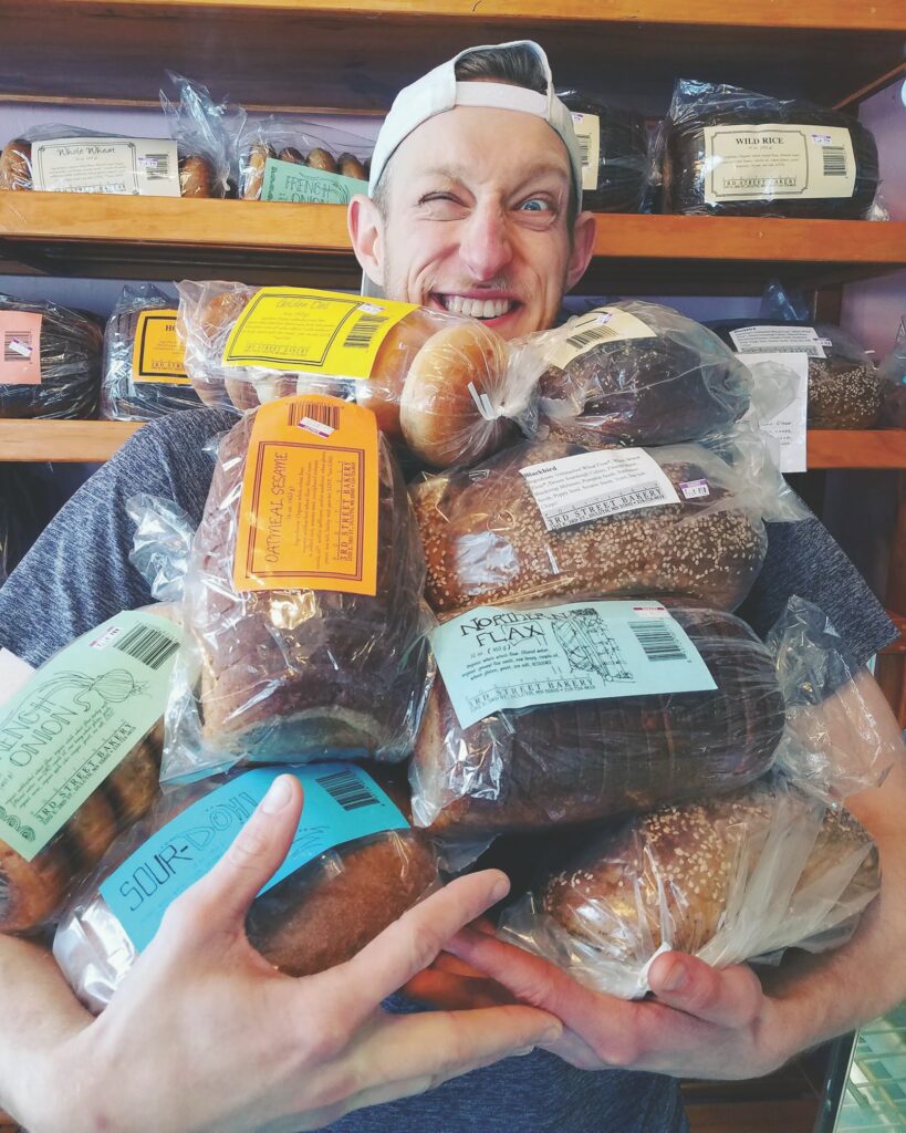 A person wearing a white cap backward is smiling while holding a large assortment of packaged bread. The background shows shelves filled with more bread.