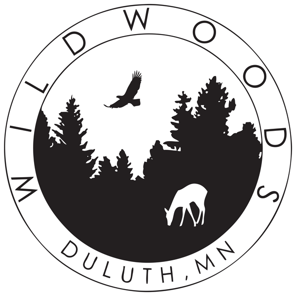 Black and white logo for Wildwoods in Duluth, MN. The logo captures a forest scene with tall trees, a deer grazing, and a bird in flight. Emphasizing wildlife preservation, the words "WILDWOODS" encircle the scene at the top, while "DULUTH, MN" is written at the bottom.
