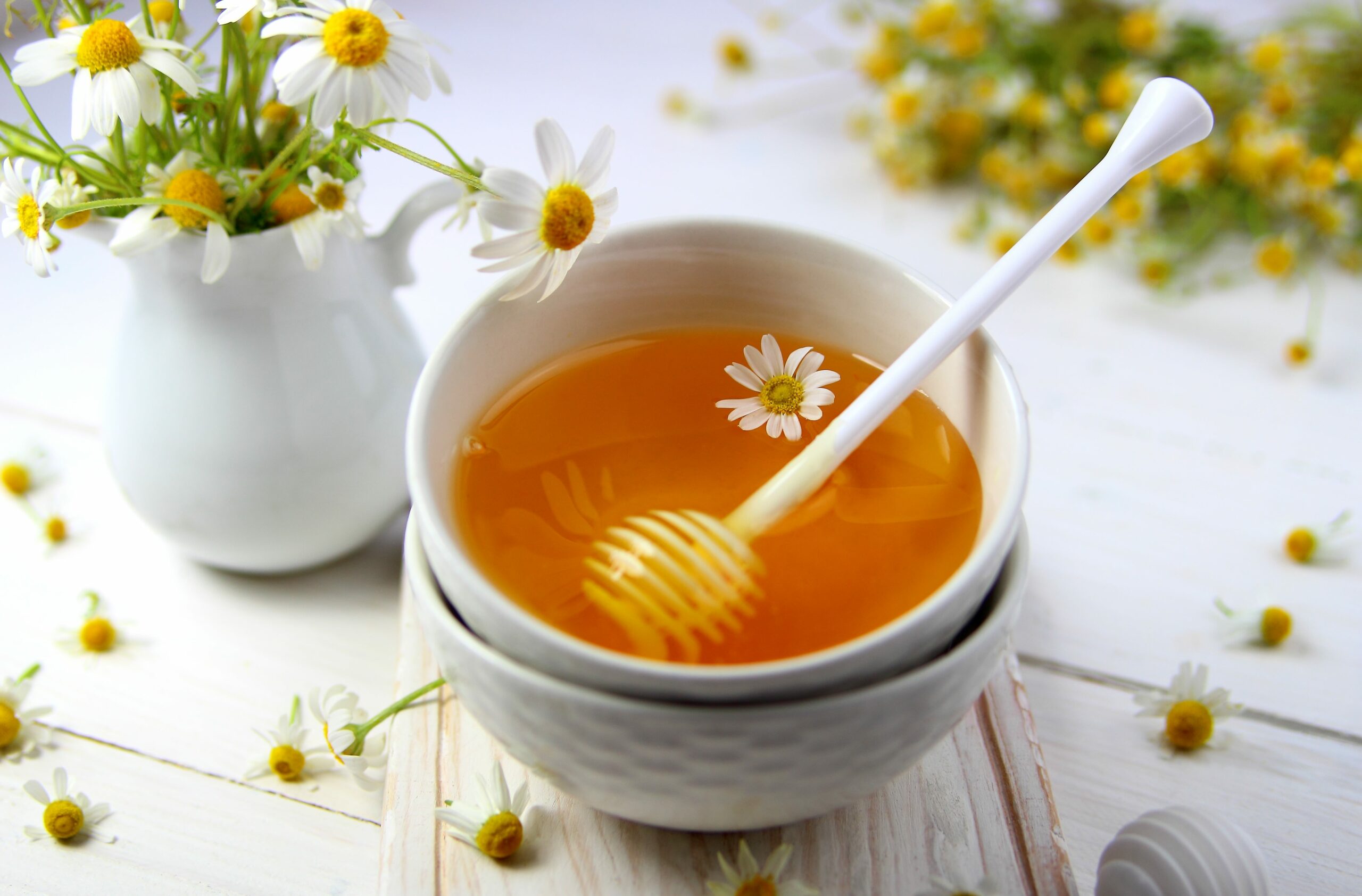 A bowl of honey with honey dipper and a vase of daisies.