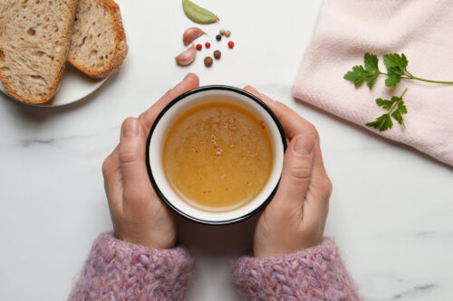 Woman with cup of hot delicious bouillon at white marble table, top view