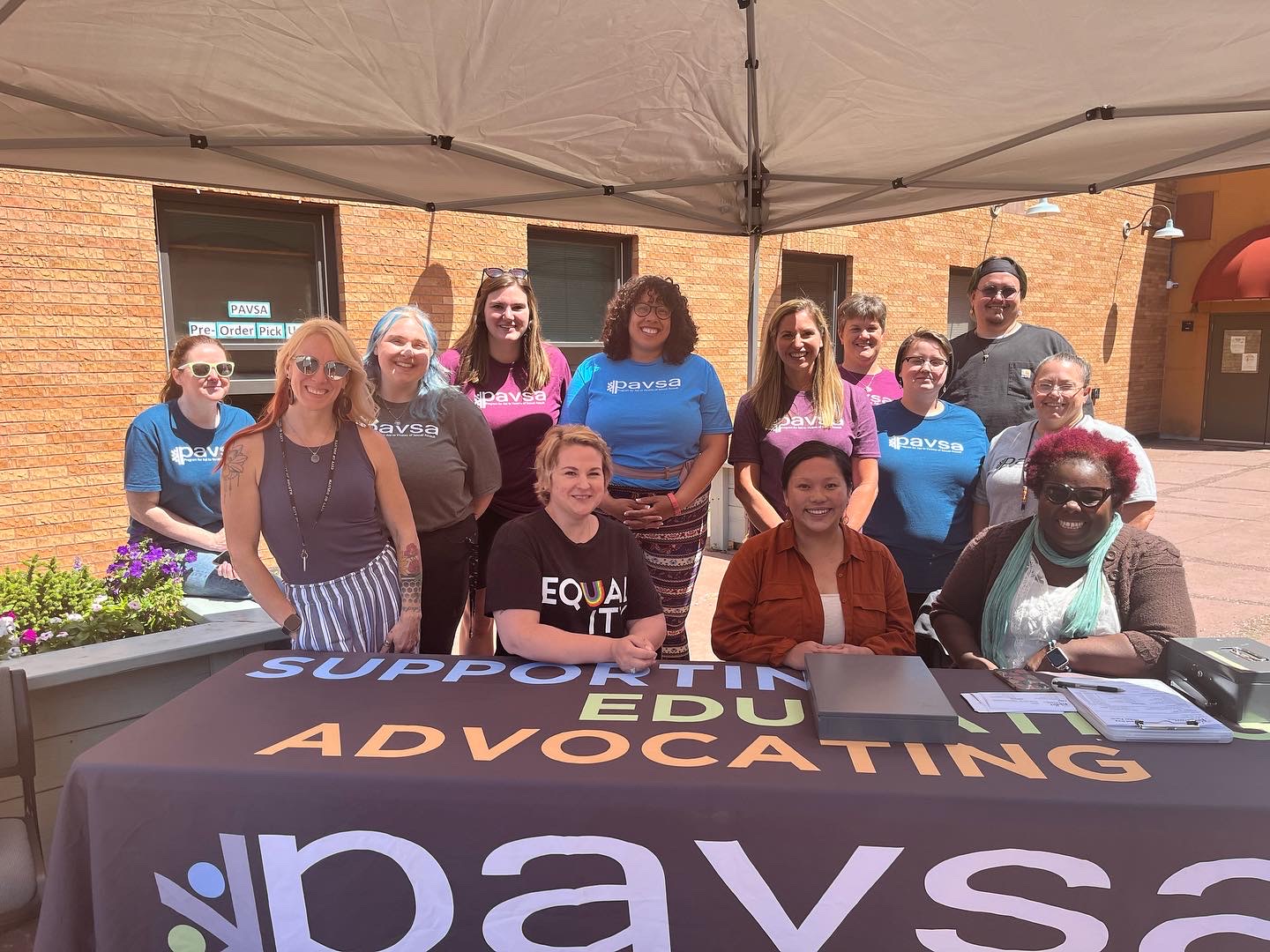 A group of people standing and sitting behind a table under a tent. The table has a sign that reads "SUPPORTING. EDUCATING. ADVOCATING." and "paysa" with a flower logo. The people are smiling and wearing various "paysa" t-shirts. The background shows a brick building.