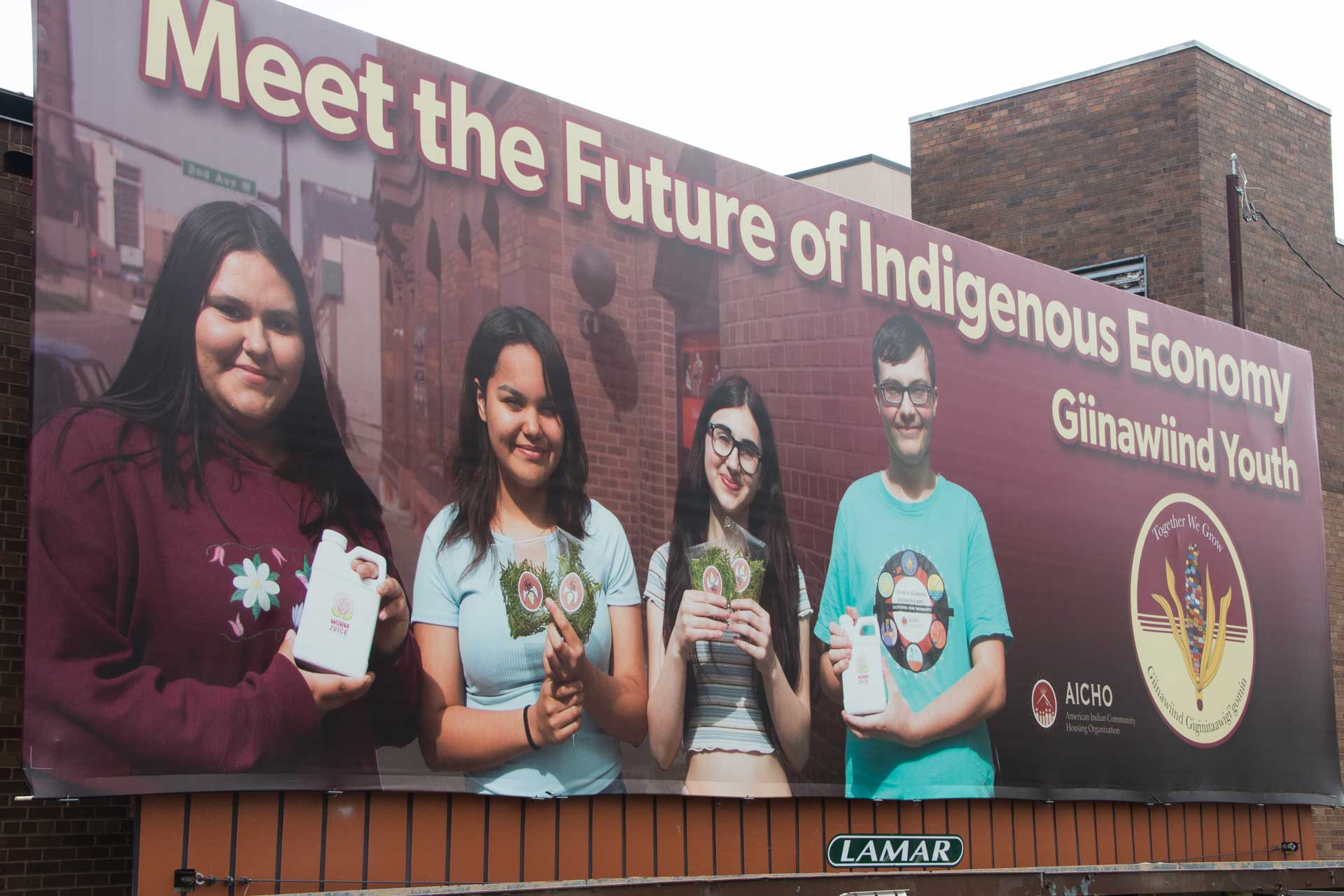 A billboard displays four smiling young individuals holding various items. The text on the billboard reads, "Meet the Future of Indigenous Economy, Giinawiind Youth" with the AICHO and Lamar logos. The background features a brick building and an urban street.