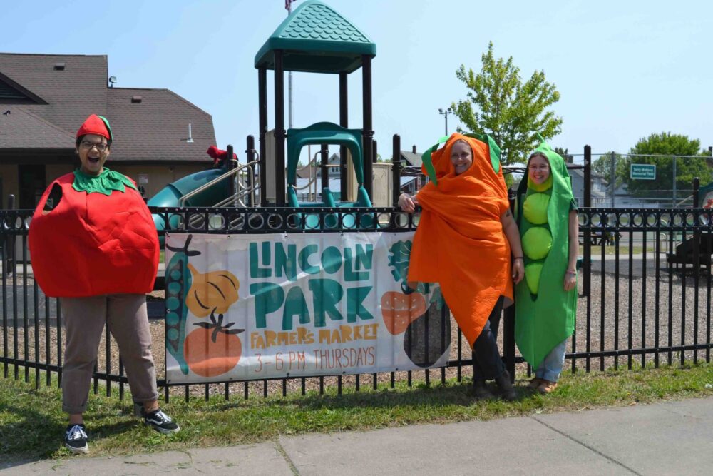 Three people wearing vegetable costumes including a tomato, carrot, and peapod stand next to a banner that says, "Lincoln Park Farmer's Market 3 – 6 pm Thursdays. There is a playground in the background.