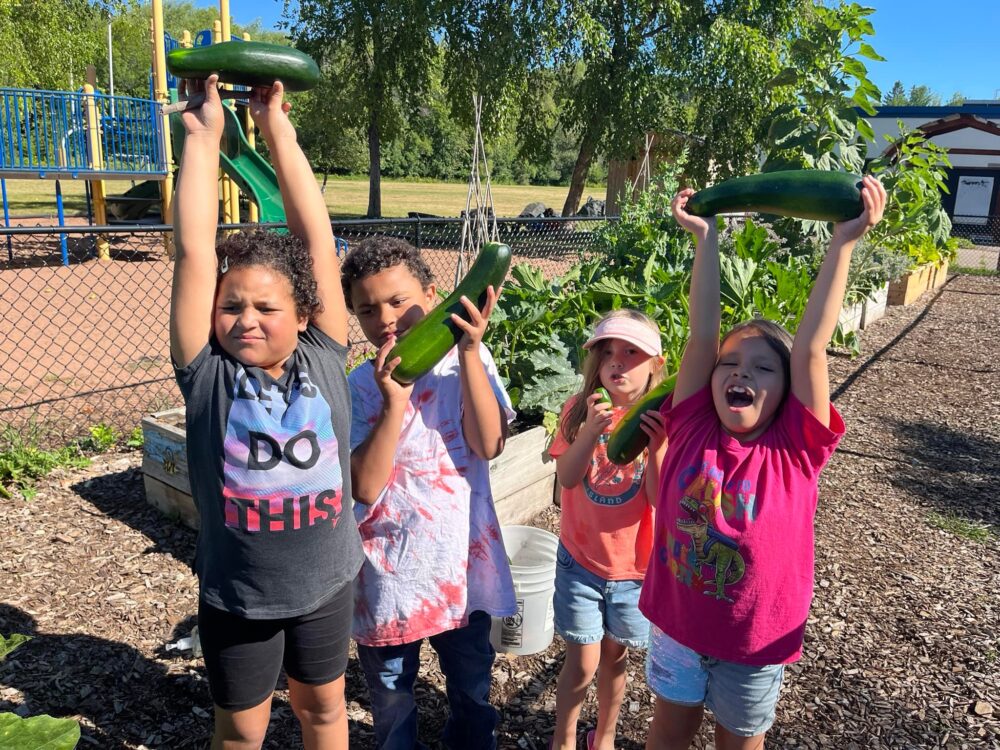 Four children each holding up a zucchini standing in front of a raised garden bed next to a playground.