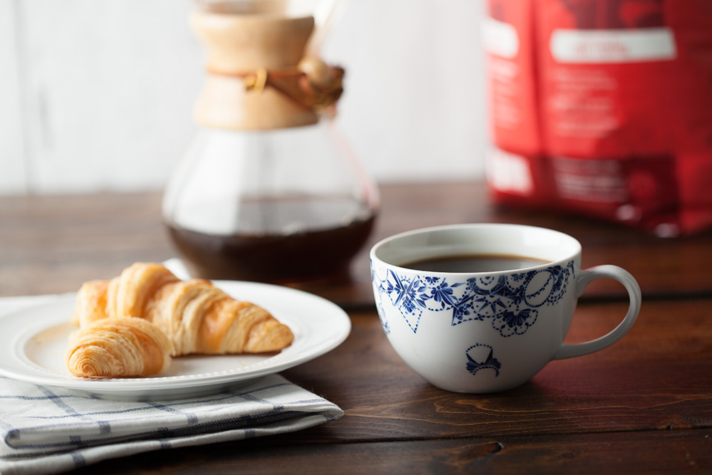 Mug of coffee next to a croissant on a plate with a Chemex and Equal Exchange coffee bag in the background