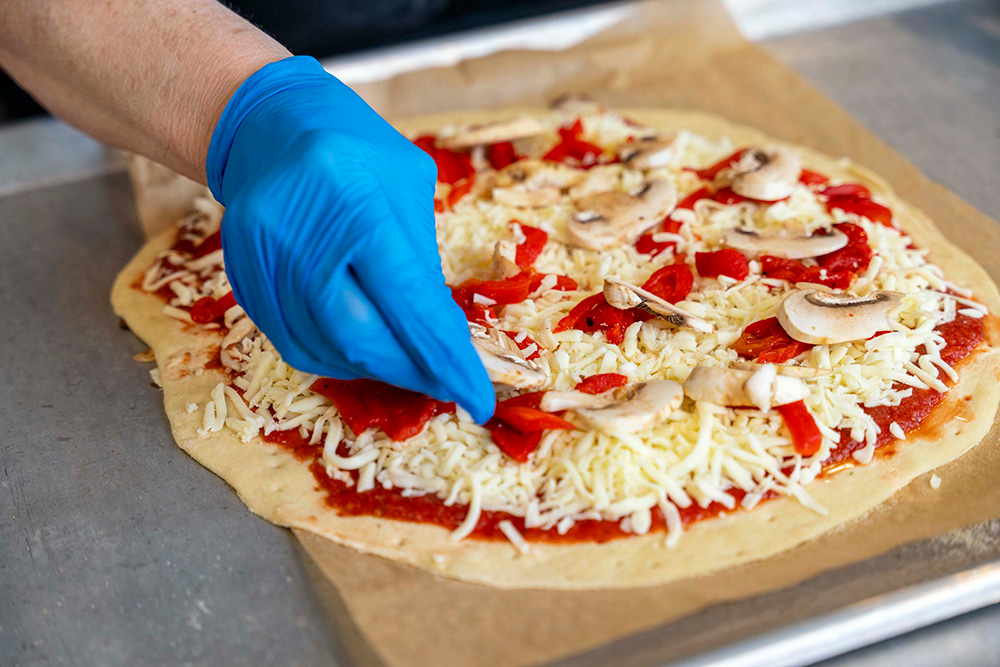 Gloved hand adding toppings to an unbaked mushroom and roasted red pepper pizza.