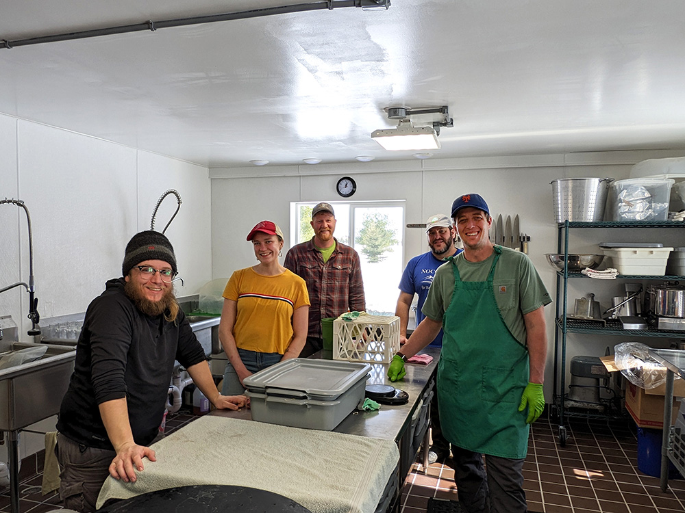 Five people stand around a prep table in a commercial kitchen.