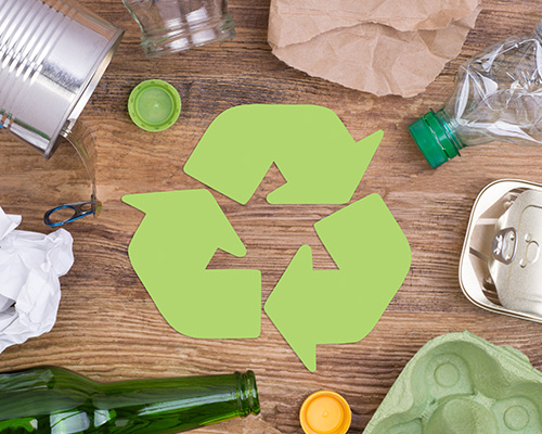 The symbol for recycling, three arrows end to end in a triangle, on a wooden table surrounded by recyclable materials including plastic bottles, glass containers, paper, an aluminum can, and an egg carton.