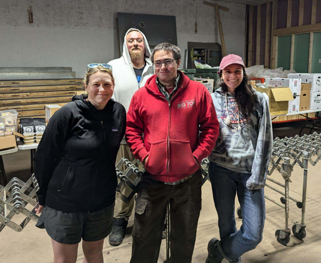 Four people pose in front on a conveyor of boxes inside a warehouse.