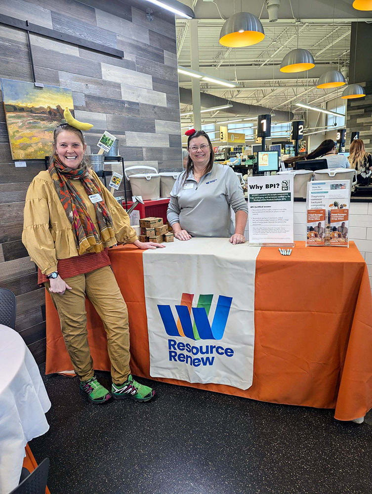 Two people at the Denfeld Whole Foods Co-op are standing next to a table with table cloth and runner with the Resource Renew logo. One is wearing a headband with a banana on top and the other has a watermelon wedge headband. There are boxes of compostable bags and information about composting on the table.