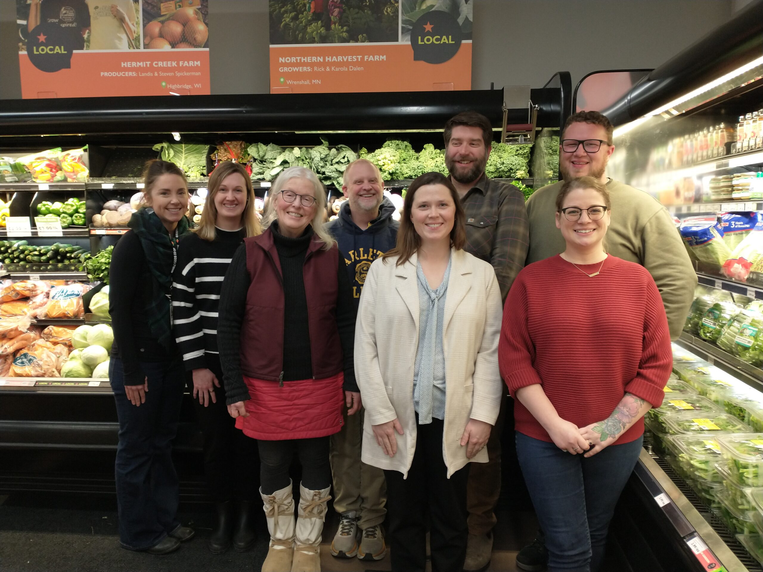 A group of eight adults smiling and posing indoors in front of a produce display at Whole Foods Co-op Hillside in Duluth, Minnesota.