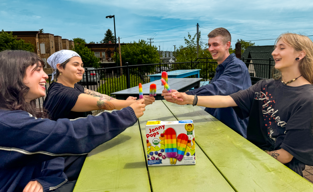 a group of young adults sitting at a green picnic table, extending their arms each holding a rainbow striped Jonny Pop frozen treat.
