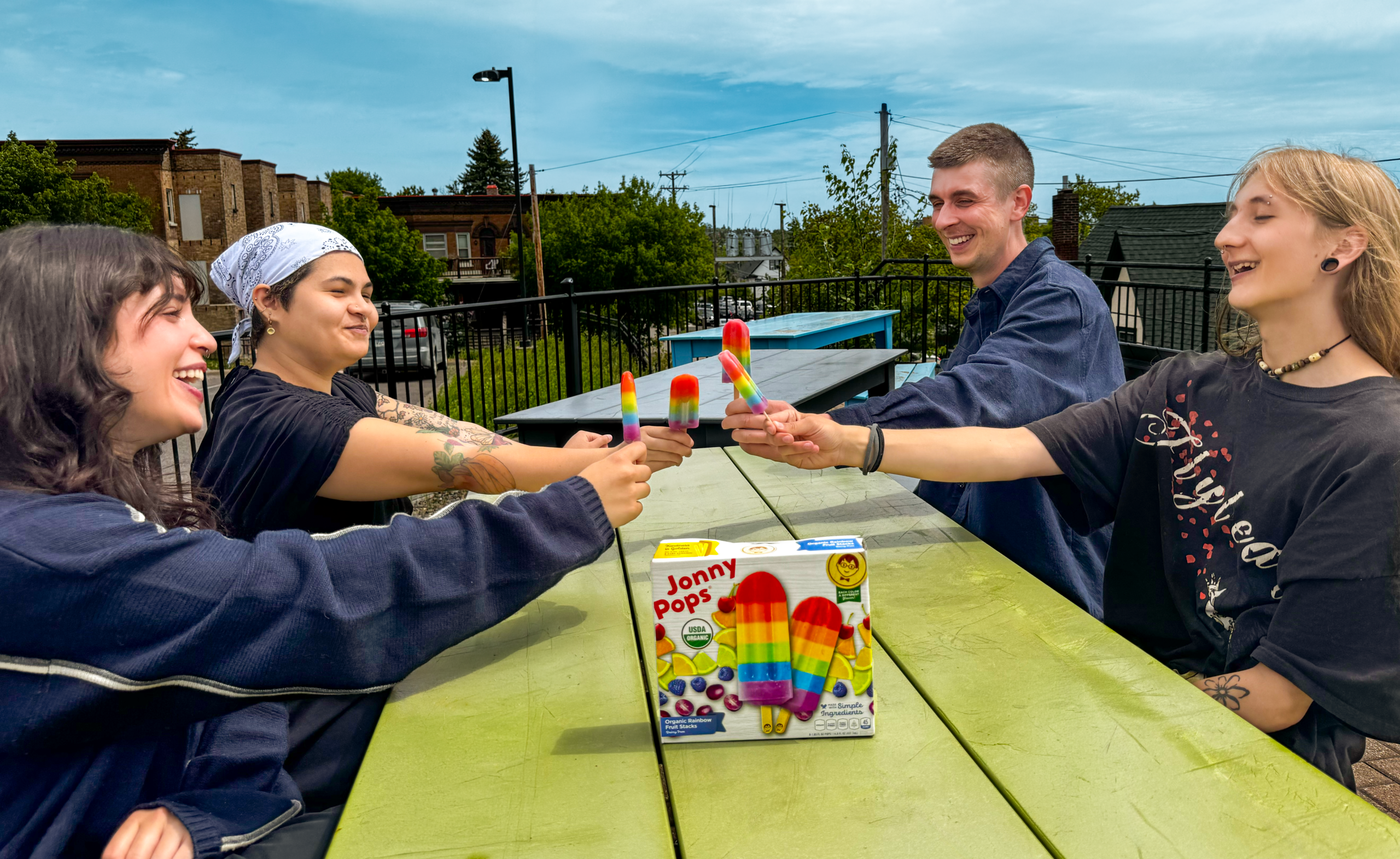 a group of young adults sitting at a green picnic table, extending their arms each holding a rainbow striped Jonny Pop frozen treat.